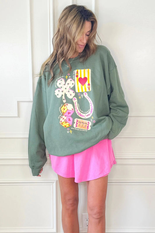 Woman wearing a green sweatshirt with colorful designs and a pink skirt against a white wall.