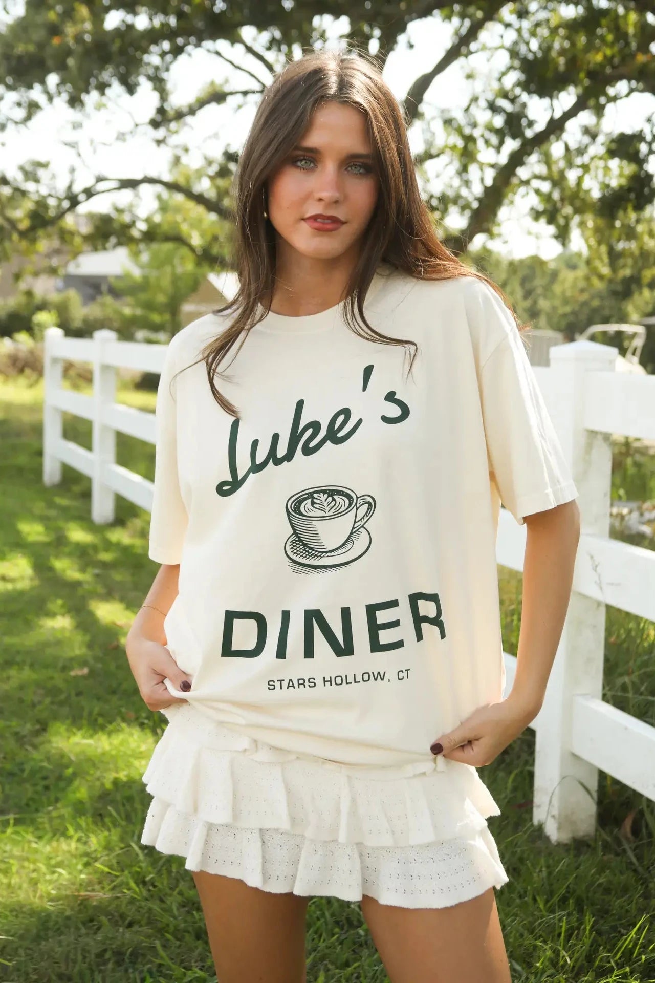 Woman wearing a 'Luke's Diner' shirt outdoors with a white fence and trees in the background