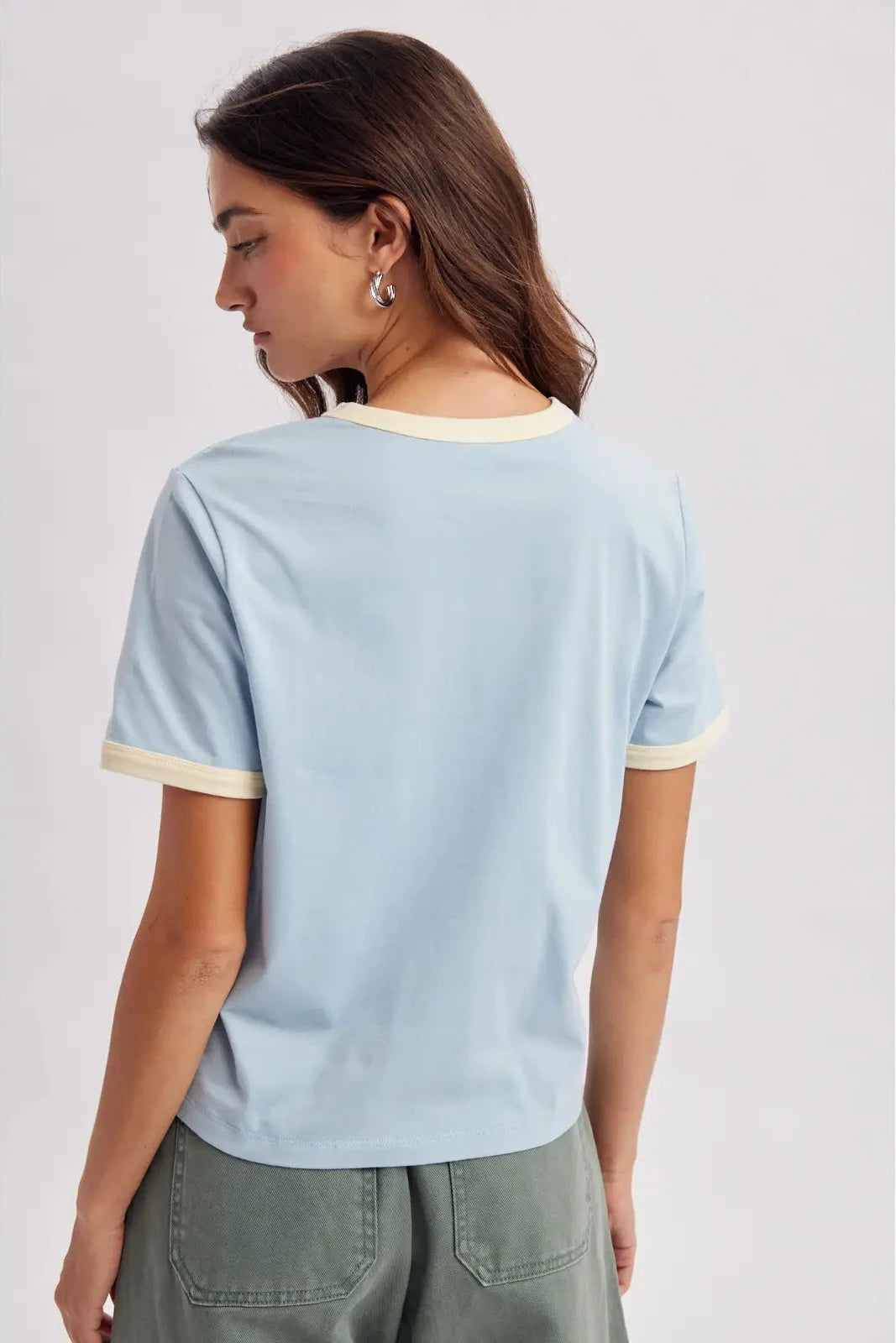 A woman shown from the back wearing a light blue ringer t-shirt with cream trim on the collar and sleeve cuffs, paired with olive green pants, against a light gray studio background.