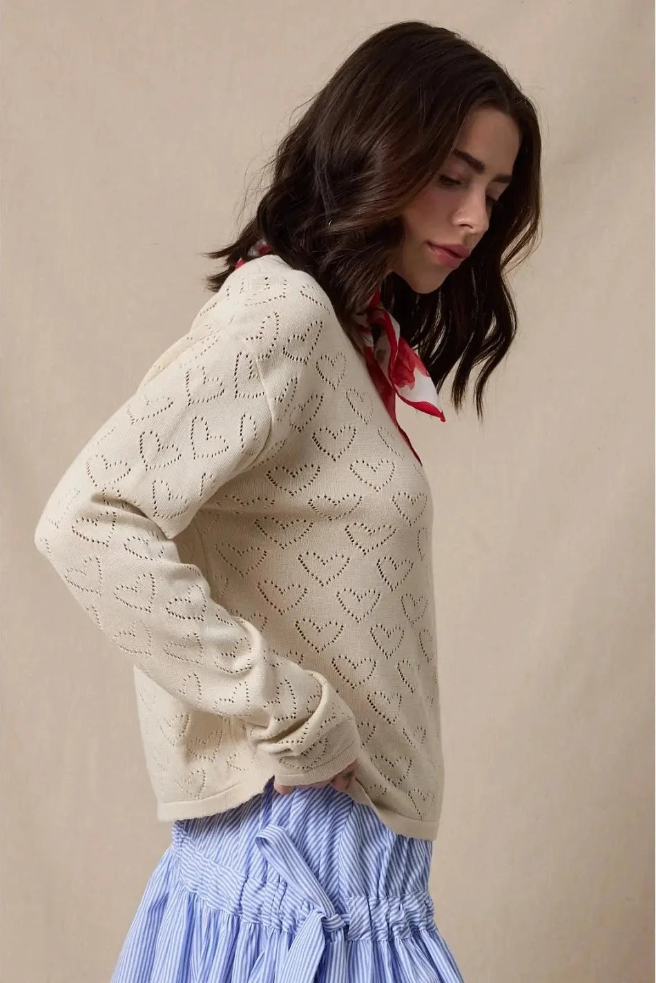 Woman wearing a cream sweater with heart patterns and a blue skirt against a beige background