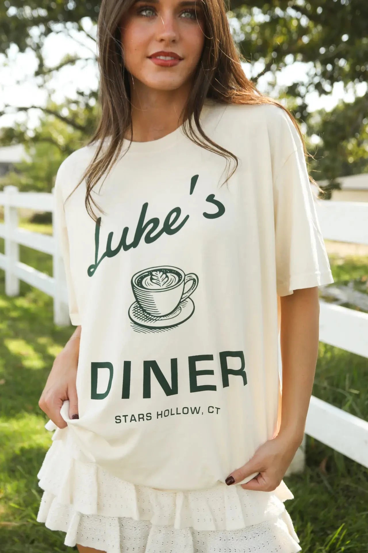 Woman wearing a 'Luke's Diner' t-shirt outdoors with a white fence and greenery in the background.