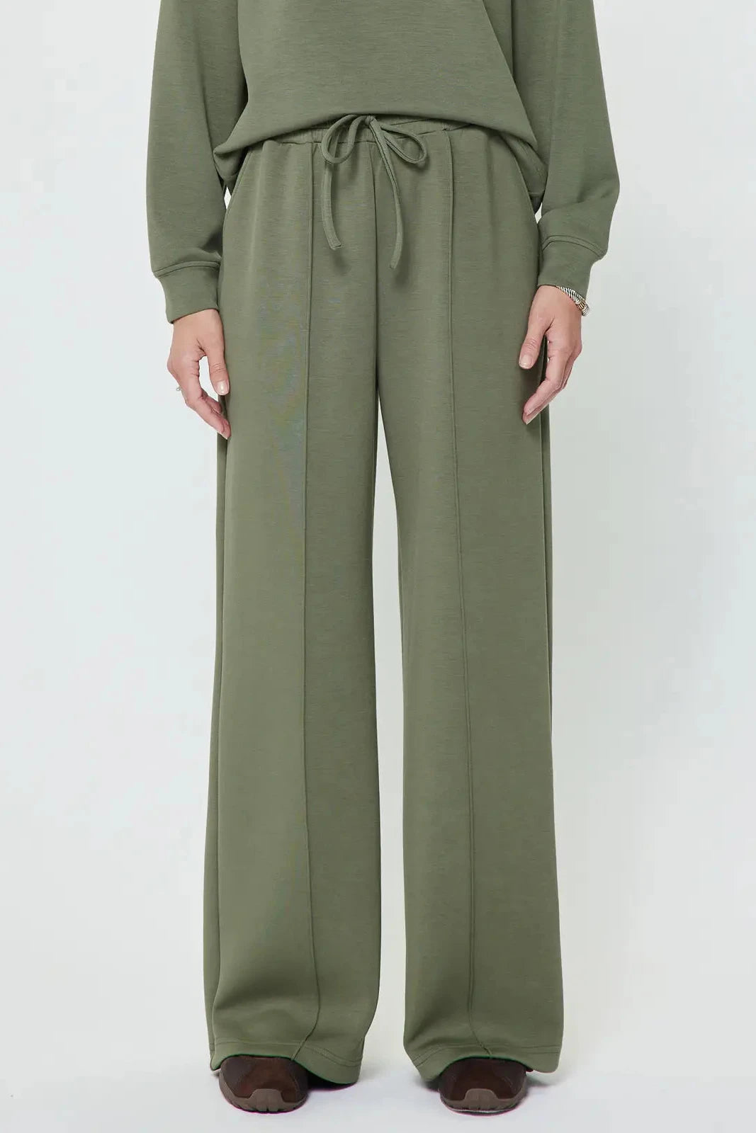 A person wearing olive green wide-leg drawstring pants with a matching long-sleeve top, shown from the chest down against a plain light gray studio background.