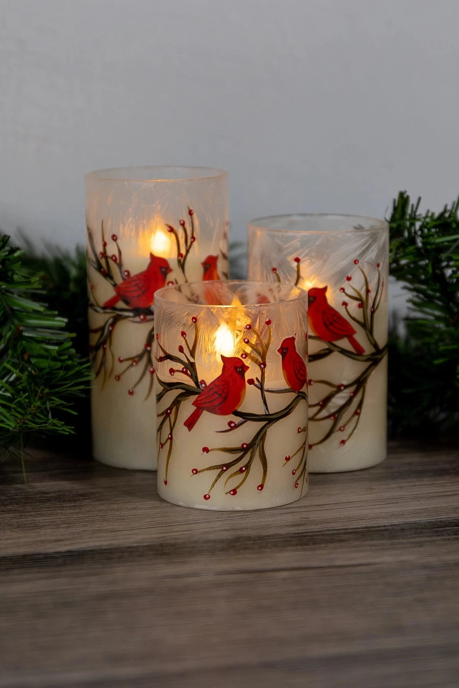 Decorative candles with red bird designs on a wooden surface with greenery in the background