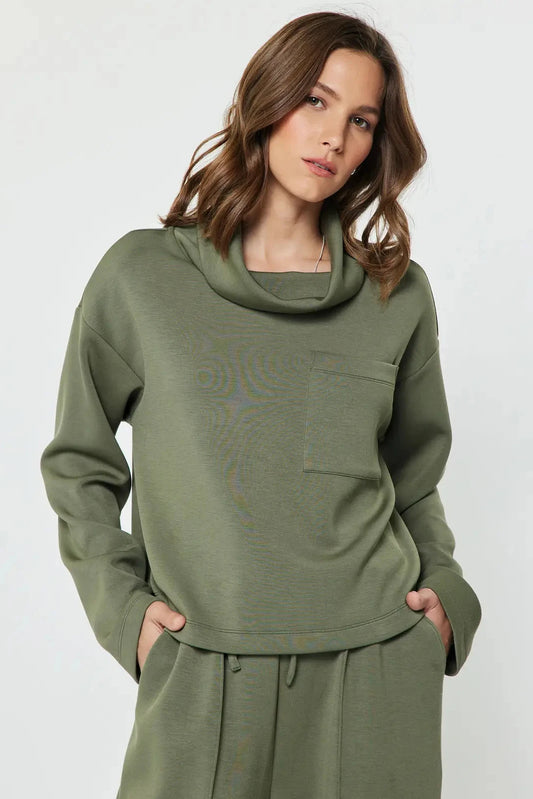 A woman modeling an olive green loungewear set featuring a cowl neck sweatshirt and matching sweatpants against a light gray studio background.