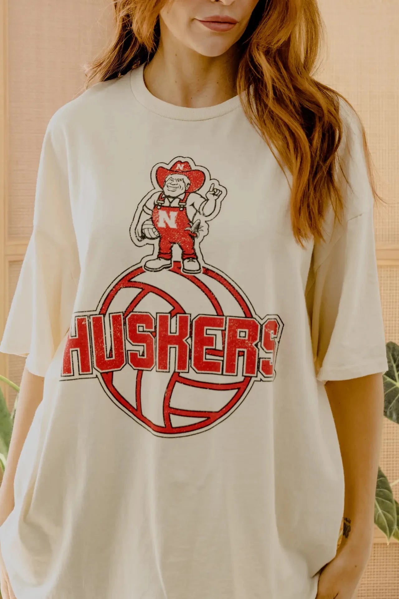 A woman wearing an oversized cream-colored t-shirt with a red and white 'HUSKERS' graphic featuring a mascot character on a volleyball.