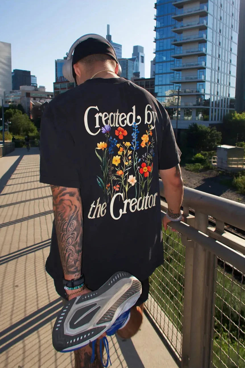Person wearing a black t-shirt with colorful floral design and text, standing on a bridge with cityscape in the background.