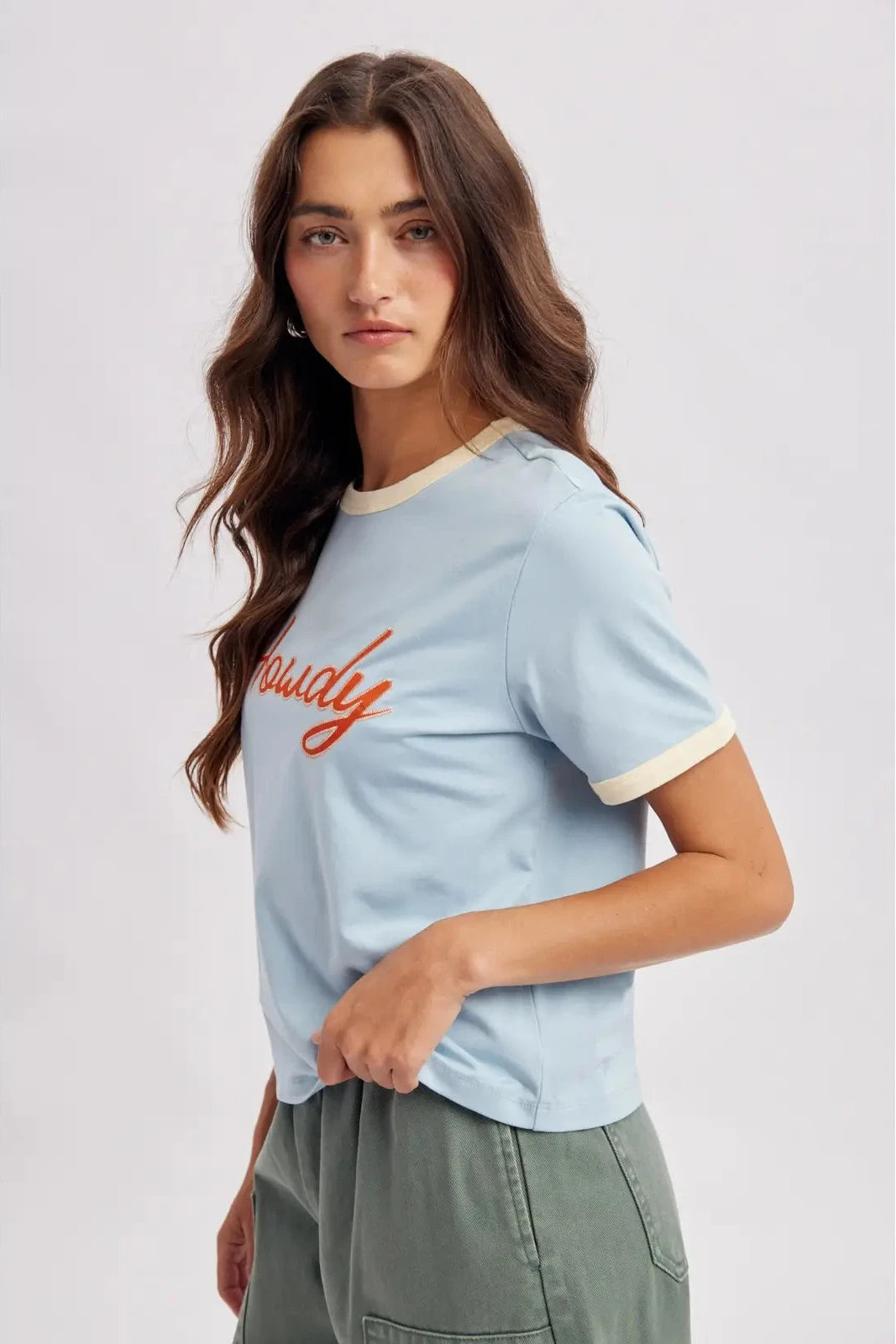 A woman wearing a light blue ringer t-shirt with cream trim and orange script text reading 'Proudly' on the chest, paired with olive green pants, against a light gray studio background.