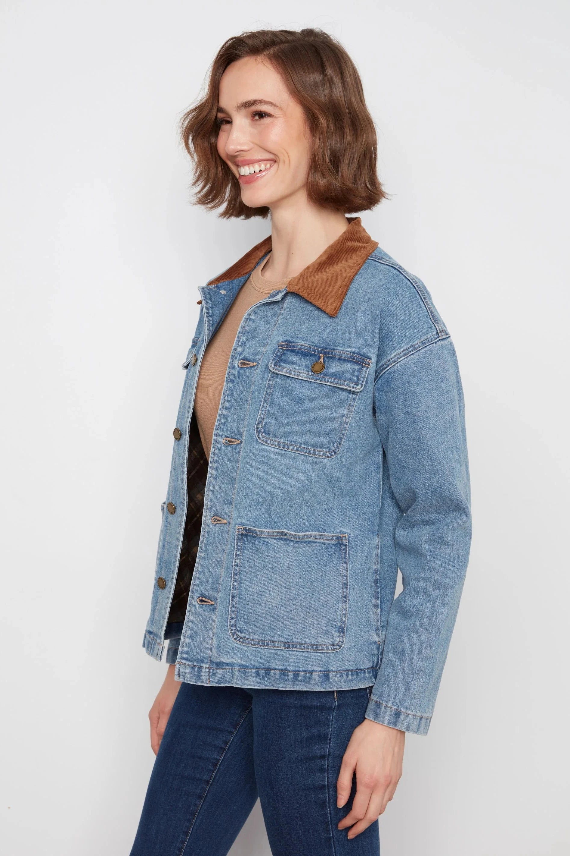 A woman wearing a light blue denim jacket with a brown suede-like collar, paired with dark blue jeans, photographed against a plain light gray studio background.