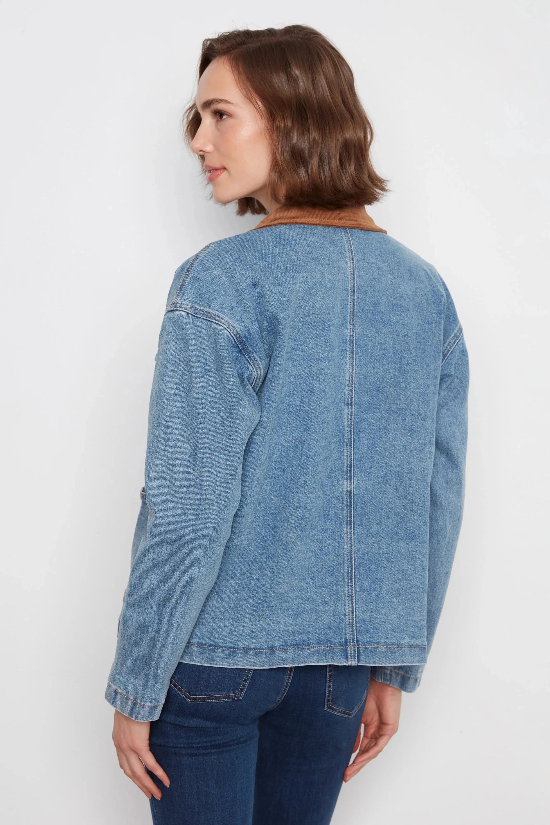 A woman shown from the back wearing a light blue denim jacket with a brown collar, paired with dark blue jeans, against a plain white background.