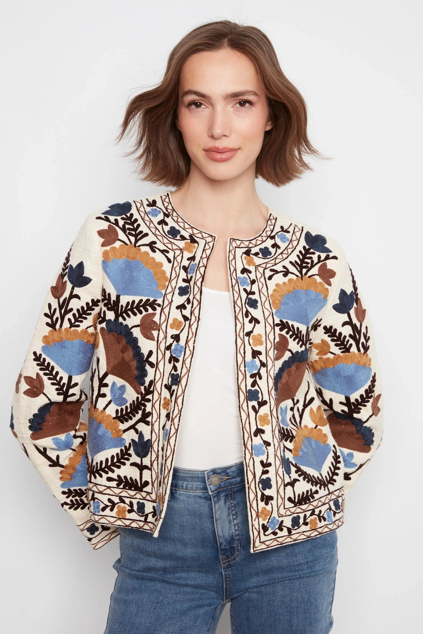 A woman wearing a cream-colored embroidered jacket with blue, brown, and black floral patterns, styled with a white top and blue jeans against a light gray background.