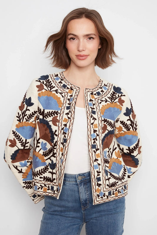 A woman wearing a cream-colored embroidered jacket with blue, brown, and black floral patterns, styled with a white top and blue jeans against a light gray background.