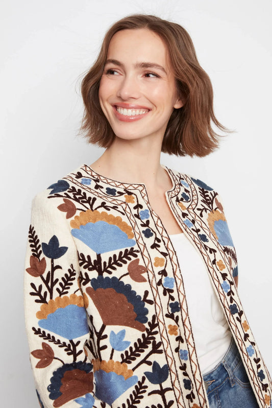 A woman with short brown hair smiling, wearing a cream-colored knit cardigan with colorful floral and leaf patterns in blue, brown, and mustard yellow, styled over a white top and blue jeans against a light gray background.