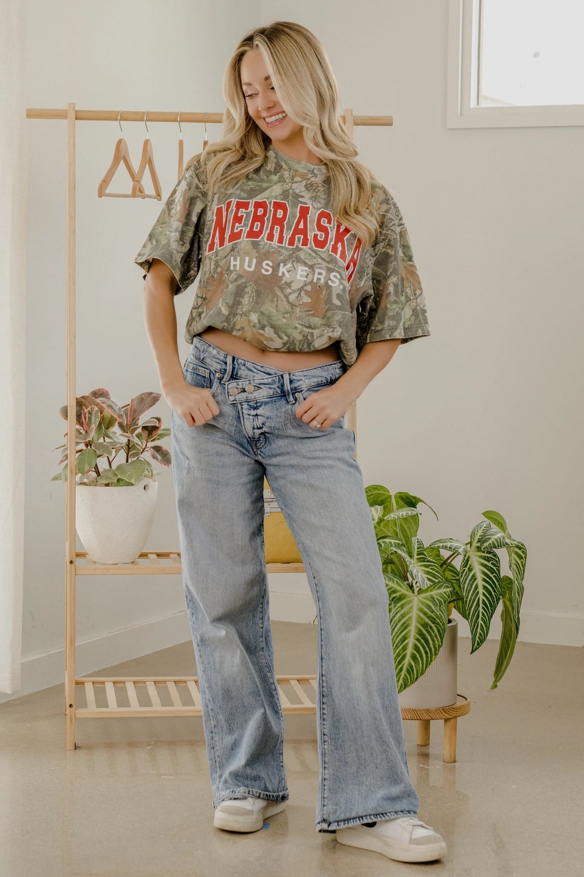 Woman wearing a Nebraska Huskers crop top and jeans in a room with plants and a rack.