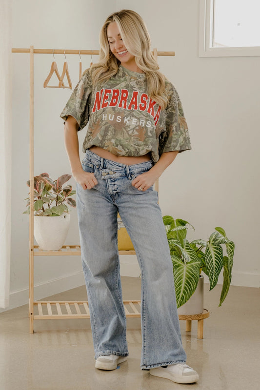 Woman wearing a Nebraska Huskers crop top and jeans in a room with plants and a rack.