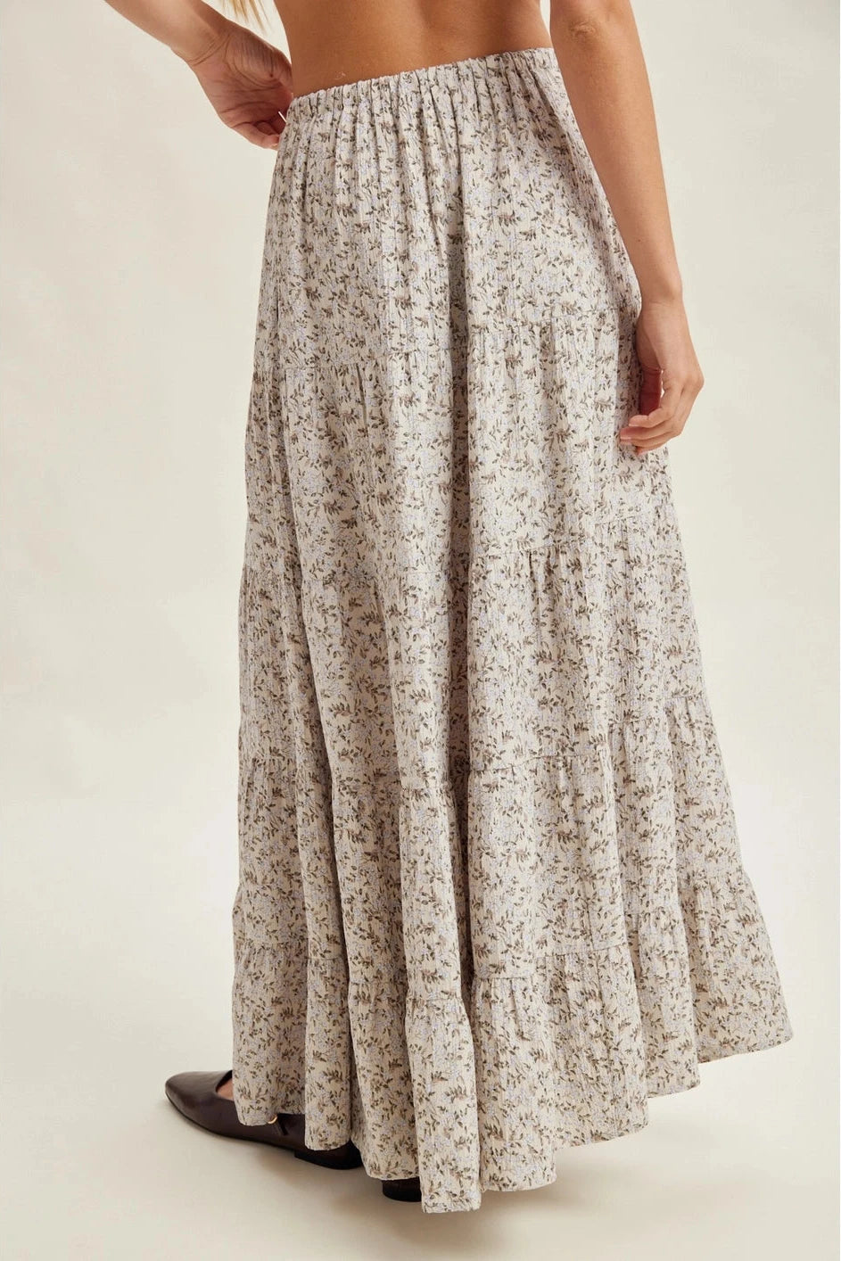 A woman wearing a long, tiered maxi skirt with a small beige and brown floral print, shown from the waist down against a plain light beige studio background.