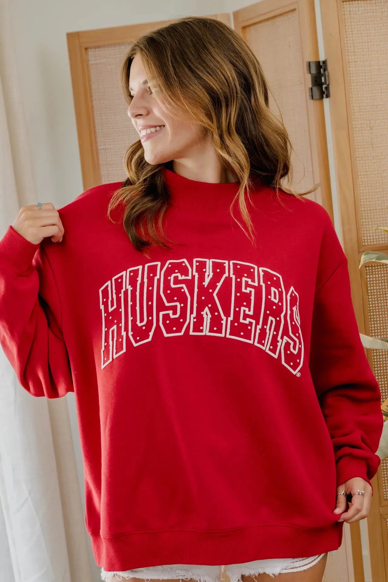 A woman wearing a red crewneck sweatshirt with 'HUSKERS' printed in white arched lettering across the chest, standing in front of a wooden screen door.