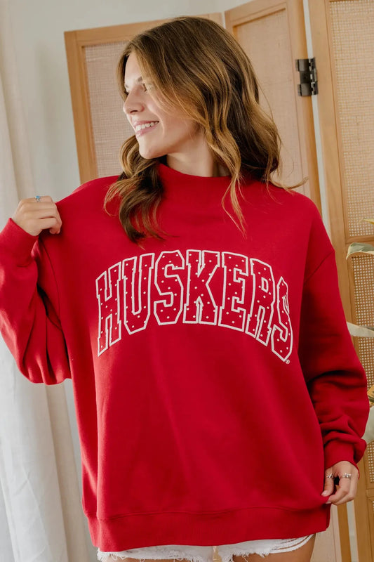 A woman wearing a red crewneck sweatshirt with 'HUSKERS' printed in white arched lettering across the chest, standing in front of a wooden screen door.