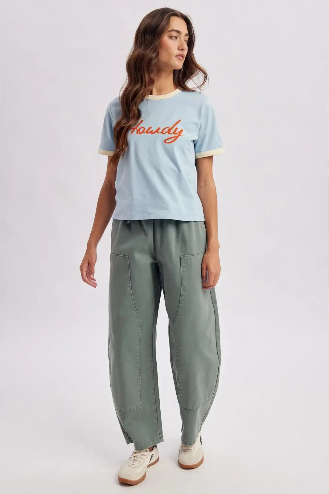 A woman modeling a light blue cropped t-shirt with orange script text reading 'Houscy' across the chest, paired with wide-leg sage green cargo pants and white sneakers, against a light gray studio background.