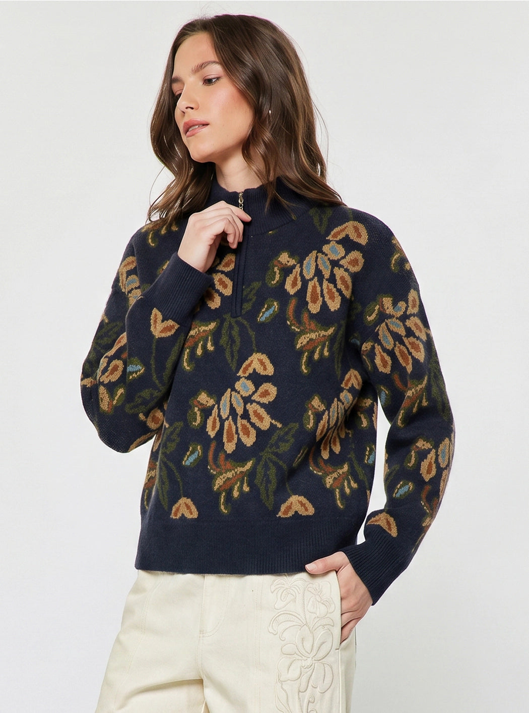A woman wearing a navy blue knit sweater with a large floral pattern in tan, olive green, and cream, paired with cream-colored pants, against a plain light gray studio background.
