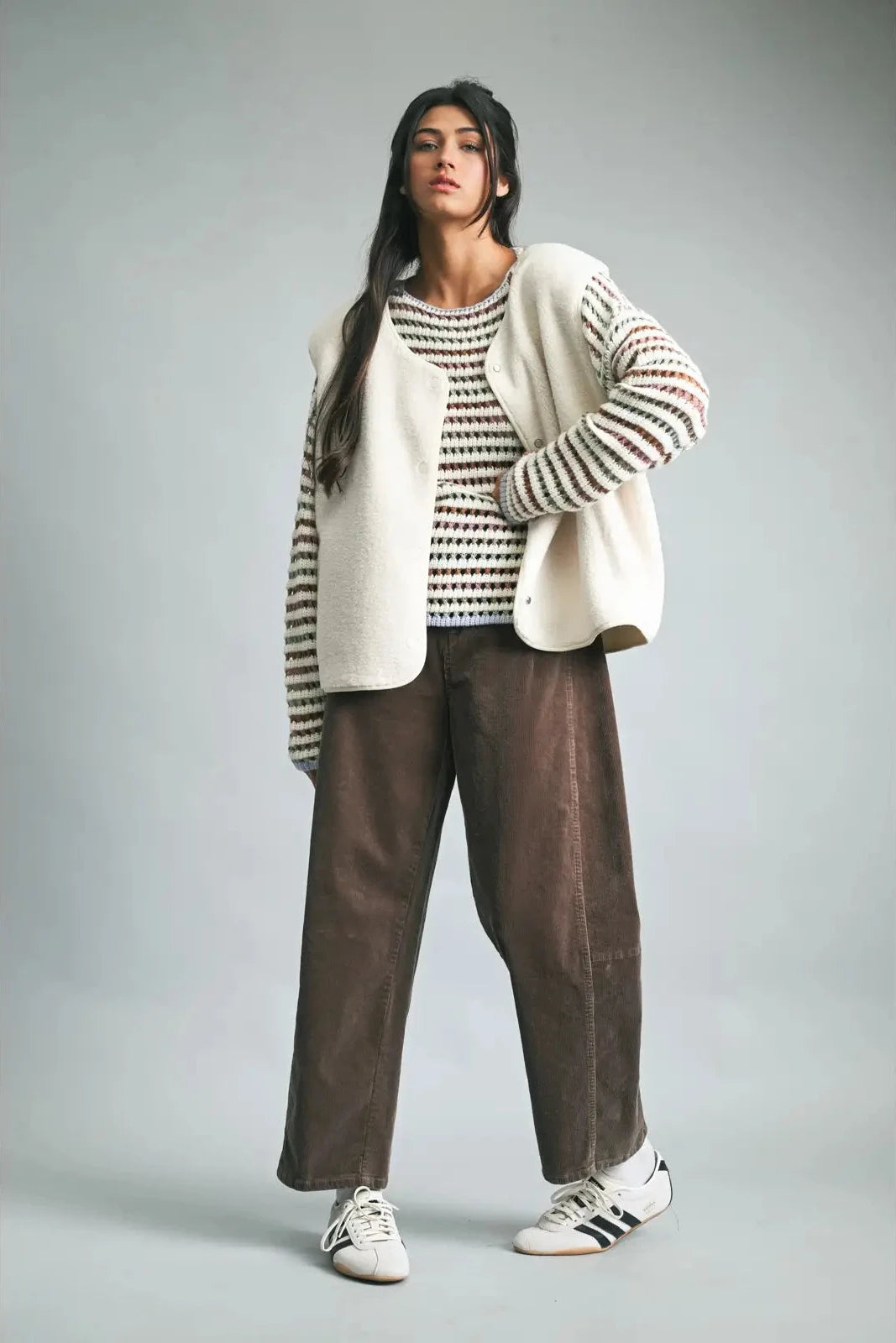 Woman wearing a white sweater with black stripes, a patterned top, brown pants, and white sneakers on a gray background