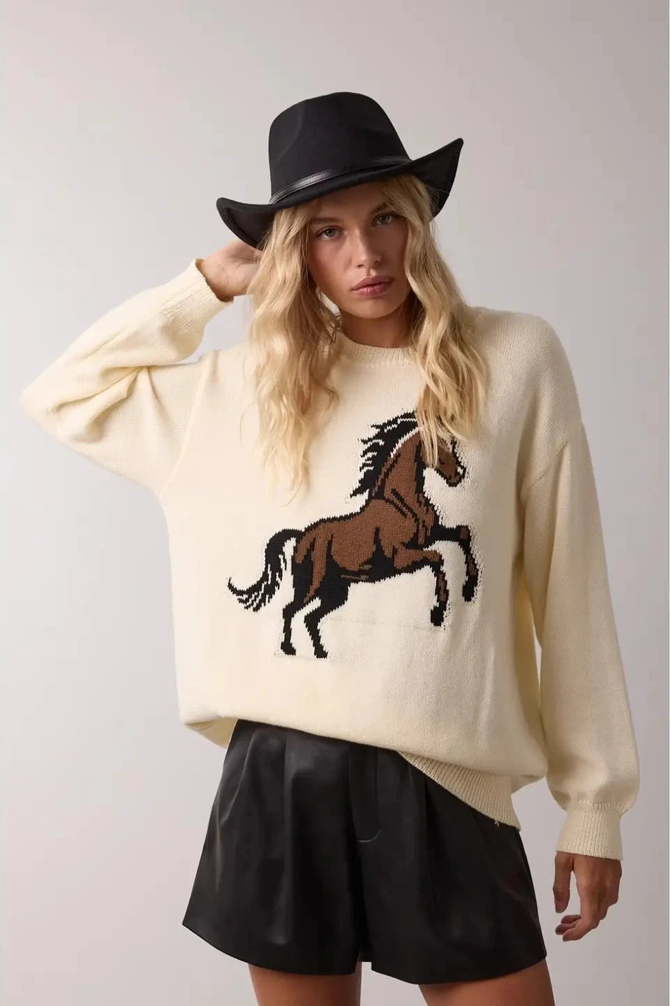 Person wearing a cream sweater with a horse design and a black hat on a plain background