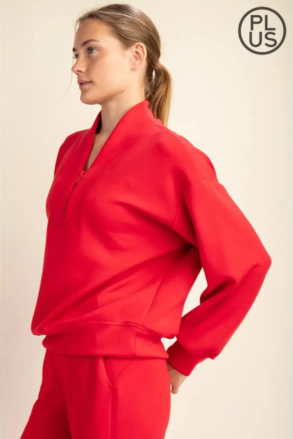 A woman wearing a bright red long-sleeve half-zip pullover and matching red pants, shown in side profile against a plain beige studio background. A black circular 'PLUS' logo appears in the top right corner.