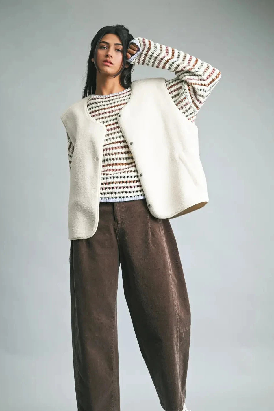 Person wearing a white vest over a patterned shirt with brown pants on a gray background