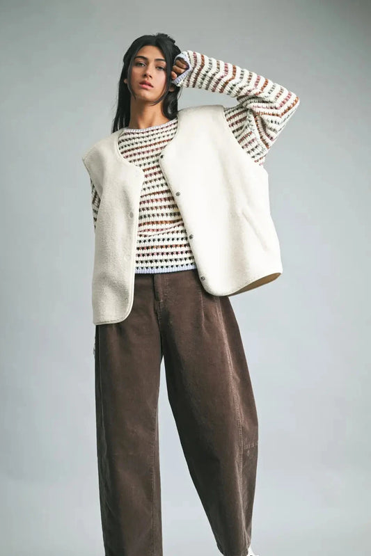 Person wearing a white vest over a patterned shirt with brown pants on a gray background