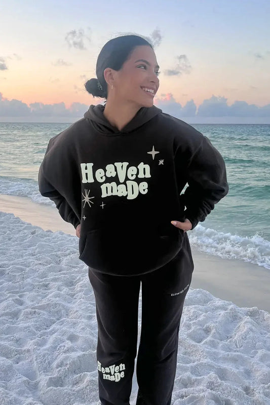 Person wearing a black 'Heaven made' hoodie and pants on a beach with ocean view.