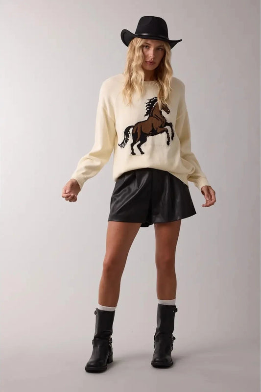 Person wearing a cream sweater with a horse design, black skirt, and black boots on a plain background