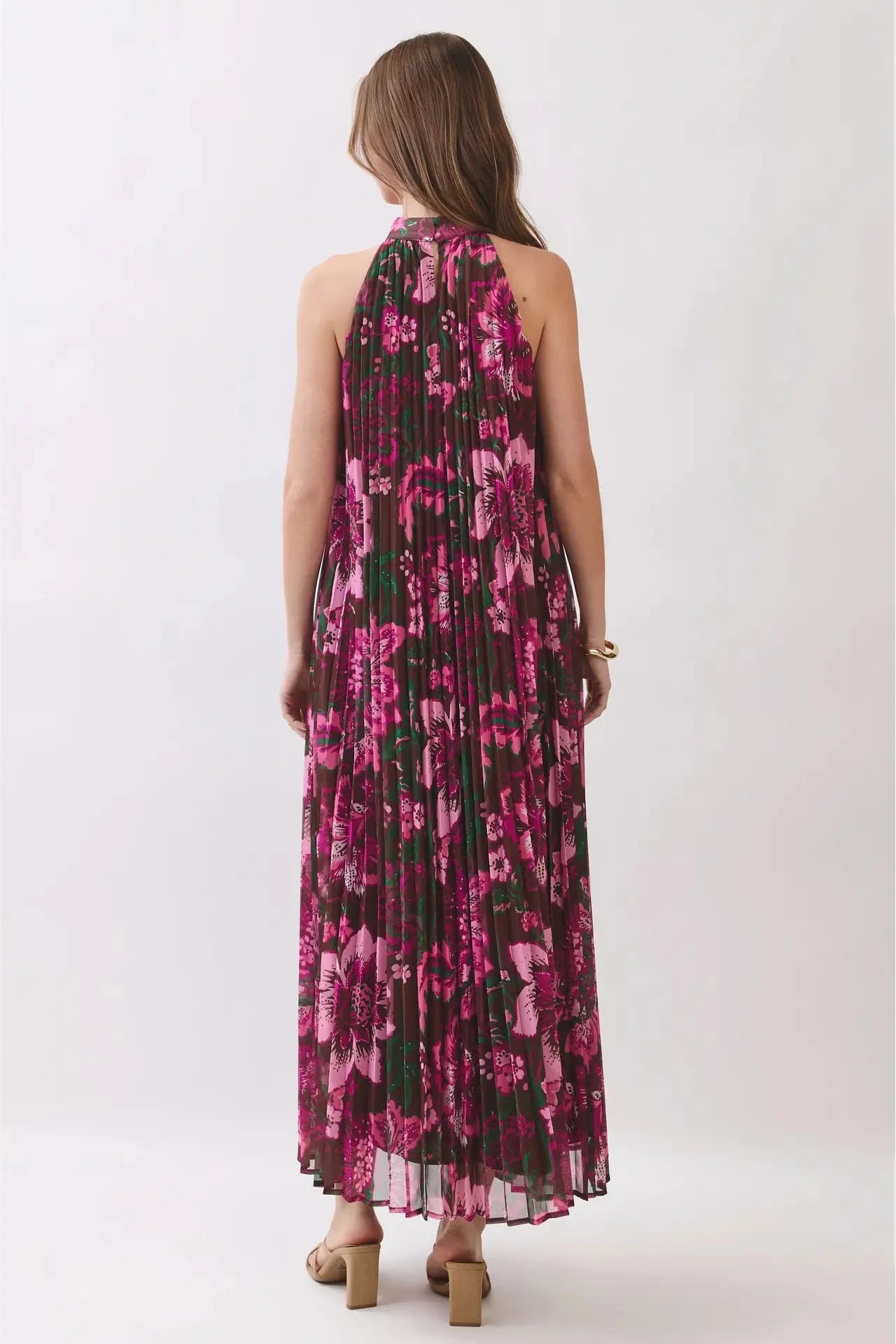 A woman viewed from the back wearing a sleeveless, high-neck, floor-length pleated maxi dress with a vibrant pink and magenta floral pattern over a black base, paired with tan block-heel sandals, set against a plain light gray studio background.