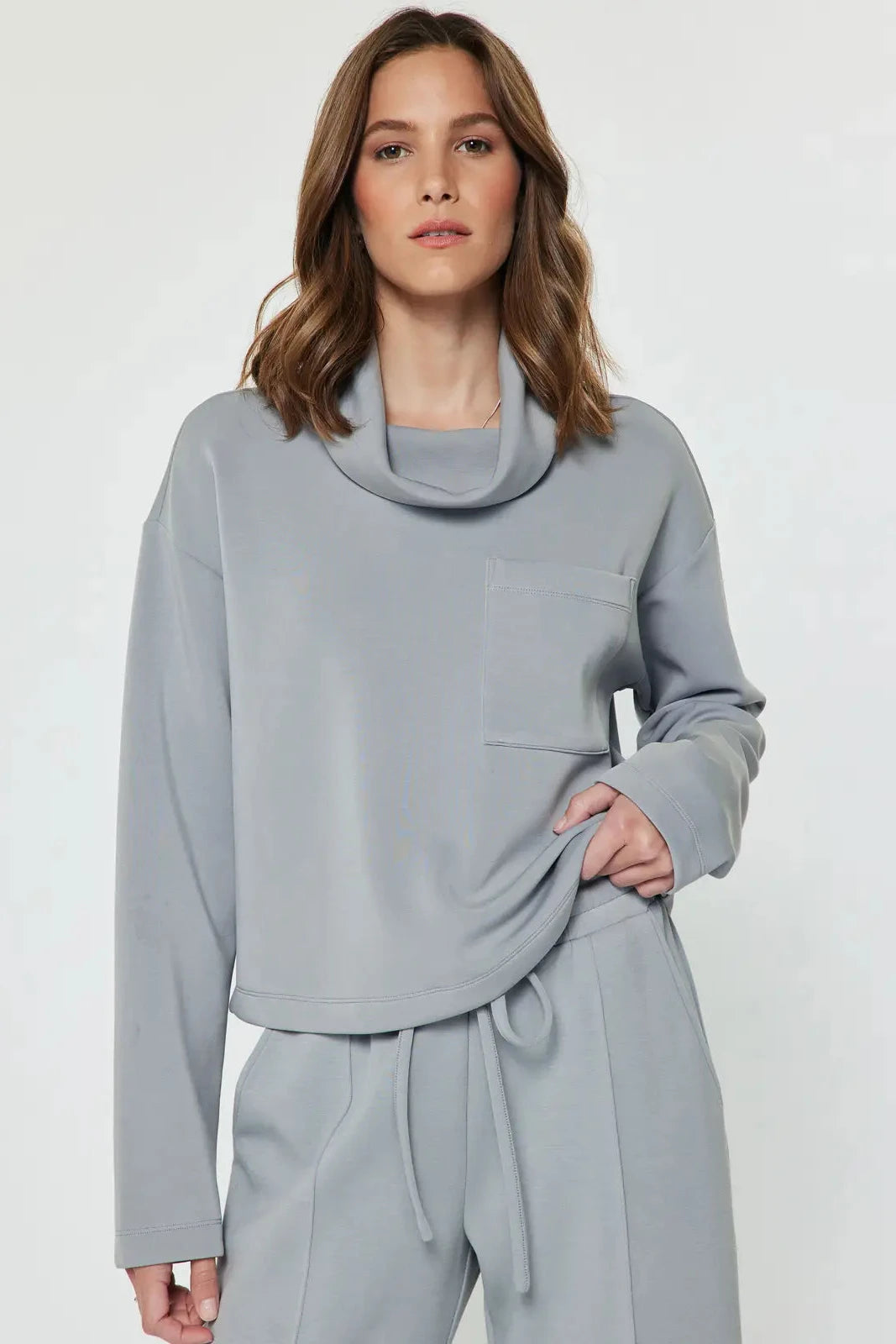 A woman modeling a light gray two-piece loungewear set featuring a cowl neck long-sleeve top and matching drawstring pants, shot against a plain light gray studio background.
