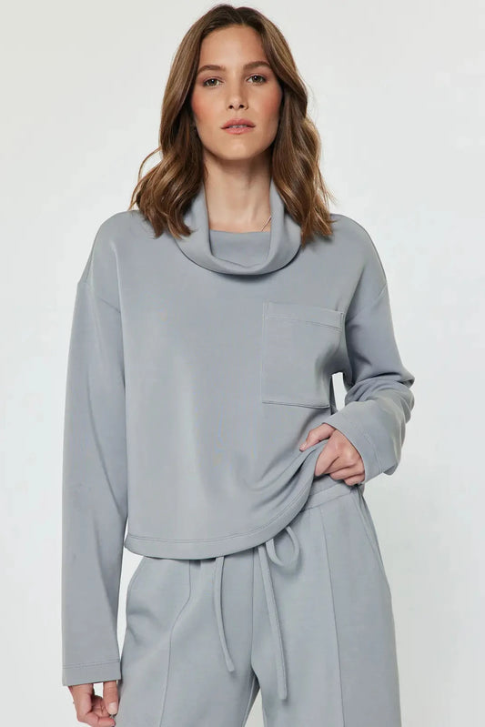 A woman modeling a light gray two-piece loungewear set featuring a cowl neck long-sleeve top and matching drawstring pants, shot against a plain light gray studio background.