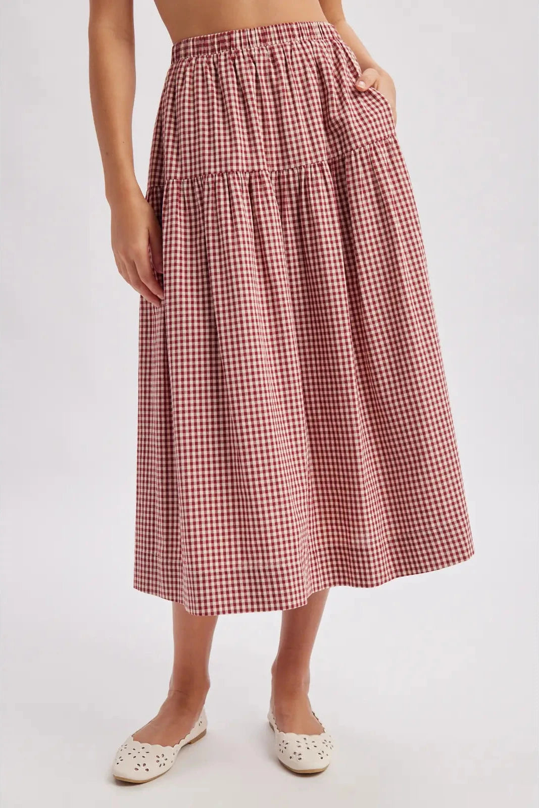 A model wearing a red and white gingham tiered midi skirt with side pockets, paired with white flat shoes, against a light gray studio background.