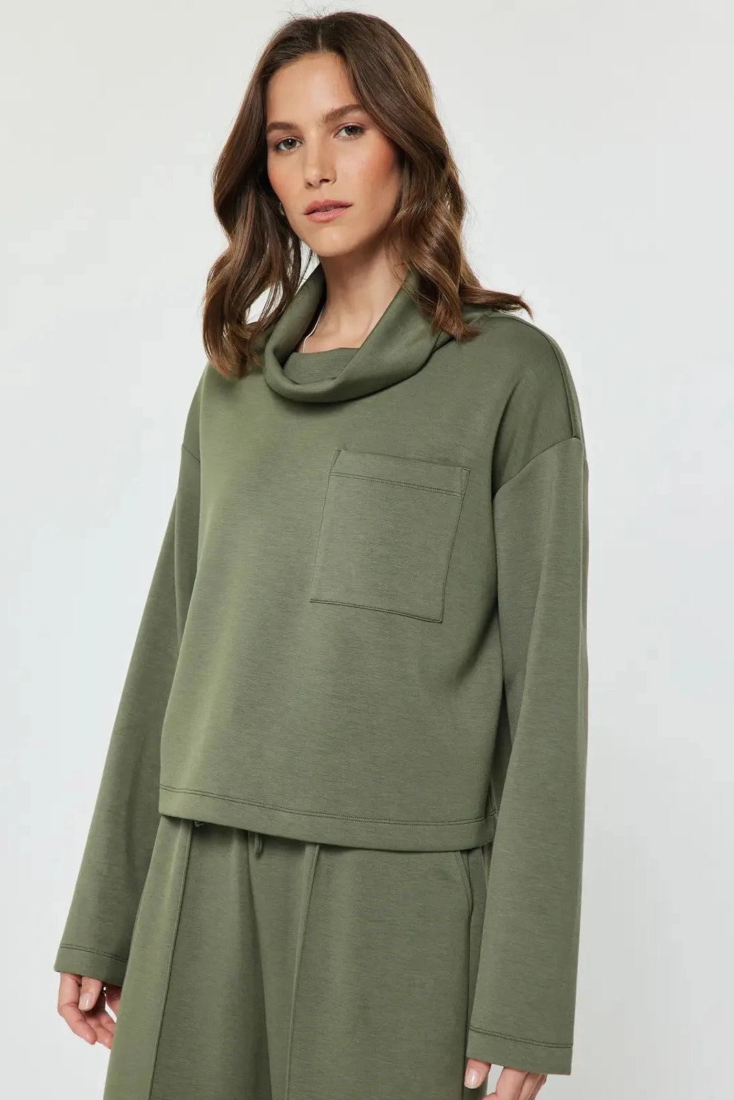 A woman wearing an olive green long-sleeve cowl-neck top with a chest pocket and matching green pants, photographed against a light gray studio background.