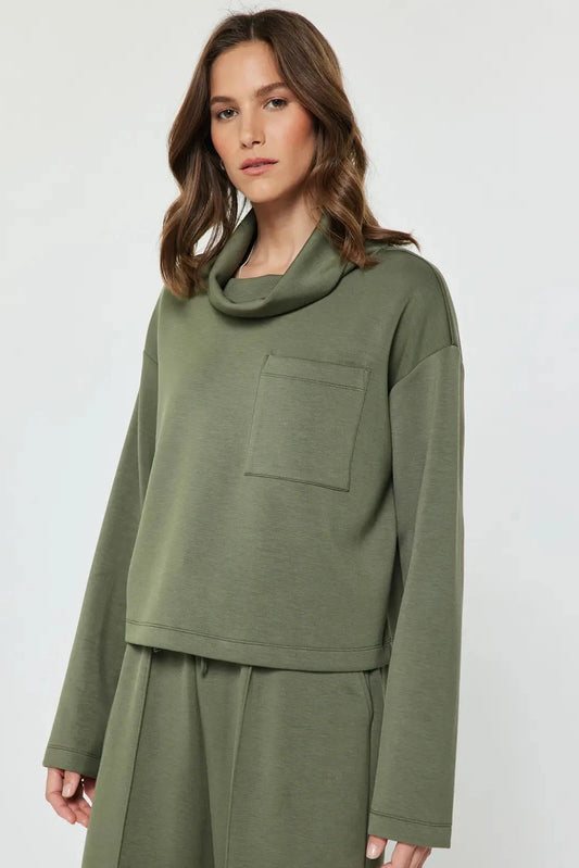 A woman wearing an olive green long-sleeve cowl-neck top with a chest pocket and matching green pants, photographed against a light gray studio background.