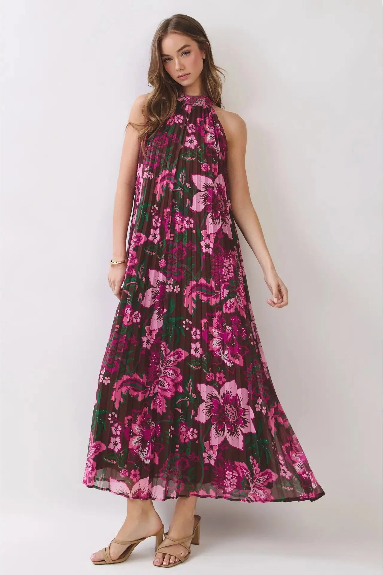 A woman modeling a sleeveless halter-neck maxi dress with a vibrant pink and magenta floral print over a dark green base, paired with tan heeled sandals against a light gray studio background.