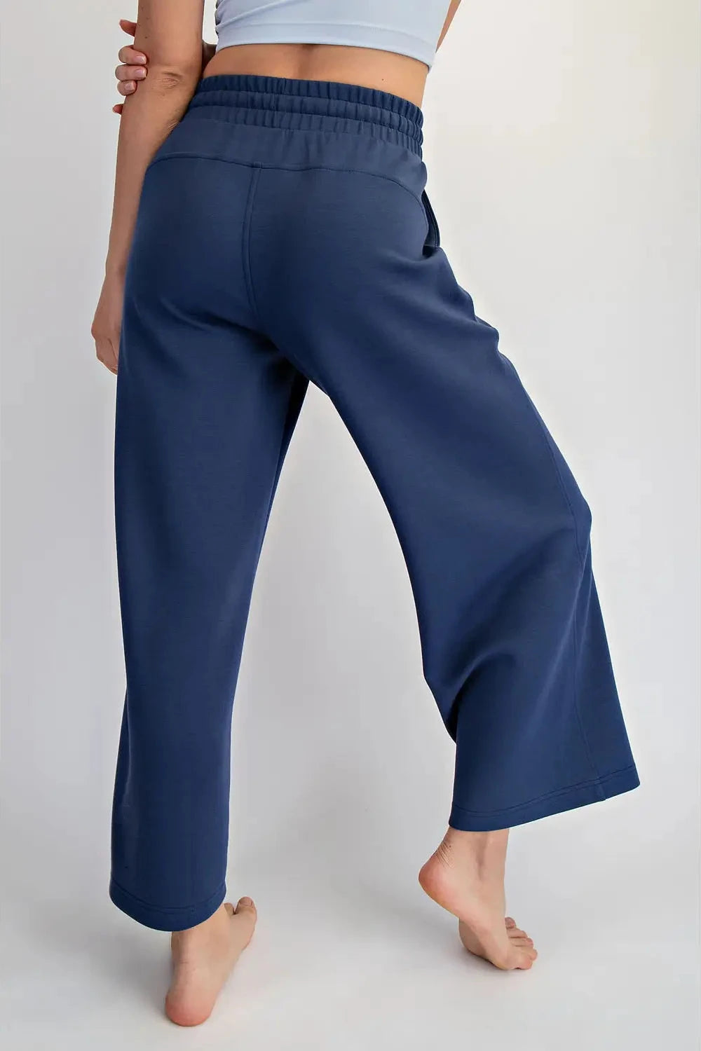 A person shown from the back wearing loose, wide-leg navy pants with an elastic waistband, standing barefoot against a plain light gray studio background.