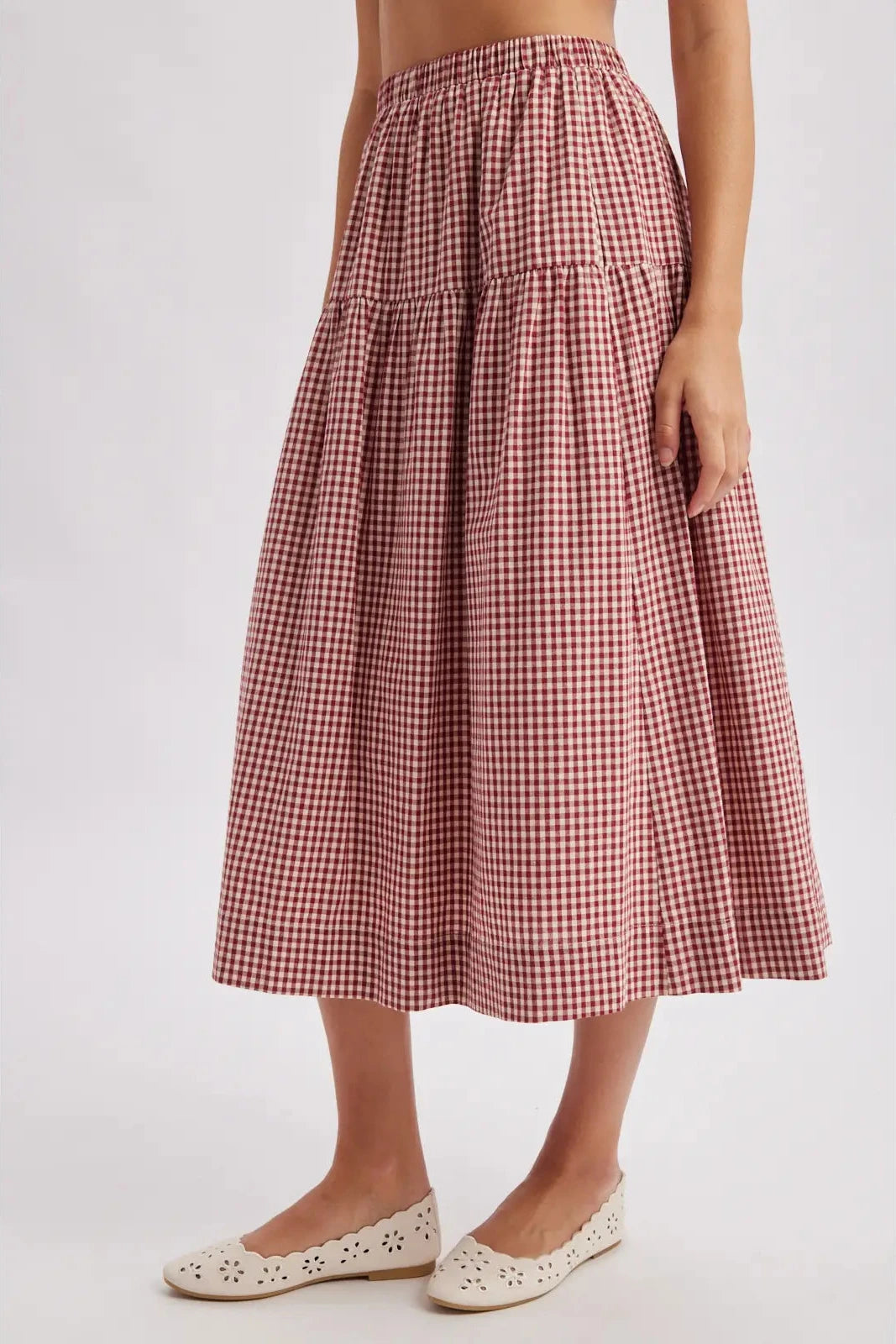 A model wearing a tiered A-line midi skirt in a red and white gingham check pattern, paired with white flat shoes, against a light gray studio background.