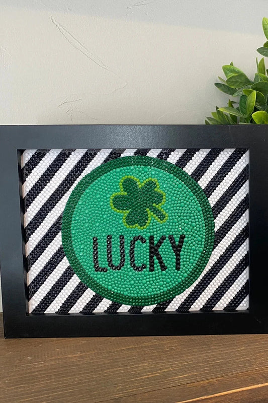 Decorative sign with shamrock and 'Lucky' text on a striped background, next to a potted plant.