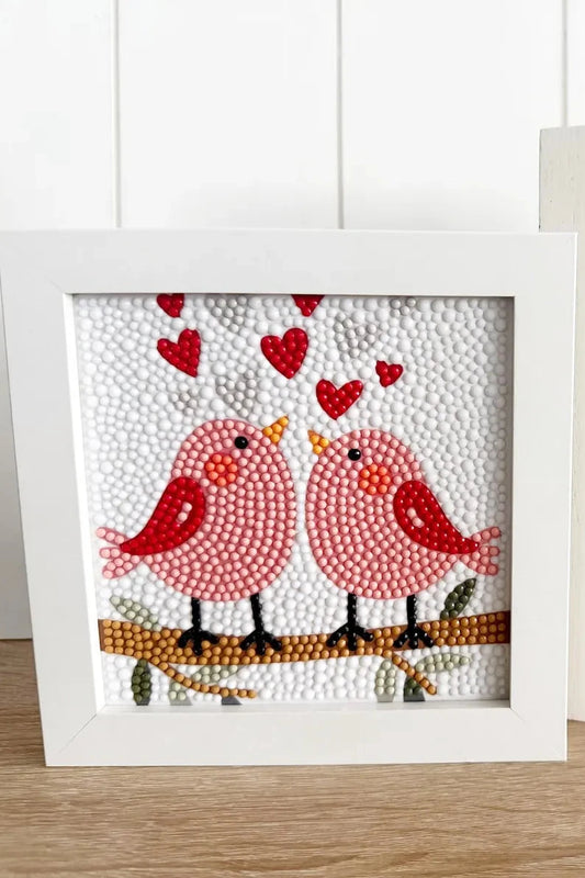 Two decorative items: a framed picture of two birds and a card with 'All You Need Is Love' text, on a wooden surface.